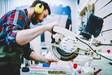 Wall Mural - serious caucasian man in headphones using circular for work with wood and timber in workshop, skille