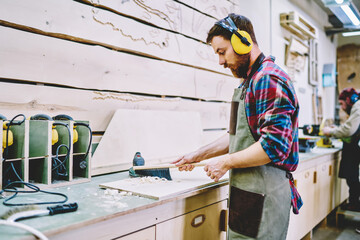 Wall Mural - caucasian male joiner in apron and headphones for safety and protection taking off sawdust after wor