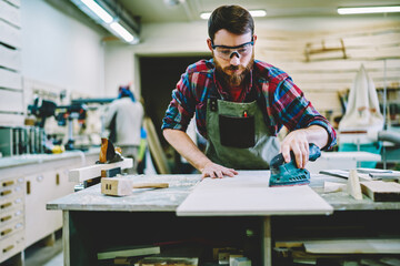 Wall Mural - serious bearded male in apron and eyewear for protection eyes while using grinder for polish timber 