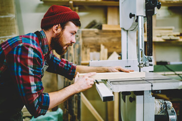 Wall Mural - serious bearded guy using modern equipment for making wooden planks in own crafts workshop, skilled 