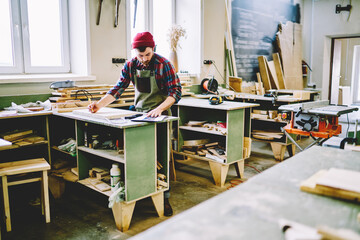 Skilled caucasian male artisan checking clients order while making furniture in manufacture workshop, serious guy looking at instructions drawing sketch on wooden plank for details on courses.