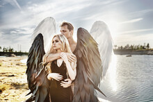 Romantic Meeting Of Angels. Good And Evil. A Parken And A Girl Stand By The Water At Sunset.