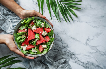 Wall Mural - Hands holding healthy fresh summer watermelon salad with arugula, spinach and greens on light marble background with tropical leaves. Healthy food, clean eating, Buddha bowl salad, top view