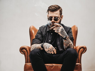 Wall Mural - Bearded tattooed man with stylish haircut in sunglasses who smokes a cigar while sitting on a vintage chair in studio