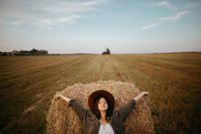 Stylish Girl Relaxing On Hay Bale In Summer Field In Sunset. Portrait Of Young Sensual Woman In Resting At Haystack, Atmospheric Tranquil Moment. Countryside Slow Life