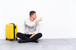 © luismolinero - Young handsome man sitting on the floor with a suitcase pointing back with the index finger