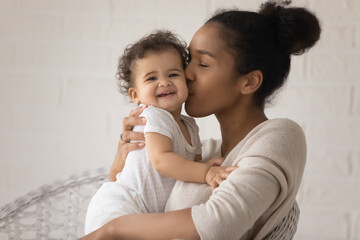 Wall Mural - close up loving young african american mother kissing cute toddler daughter cheek, sitting in cozy a