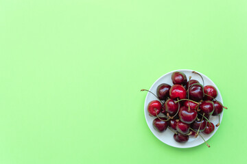 Fresh sweet cherries on white plate on green background. Healthy eating, minimalism concepts. Flat lay style with copy space for your text.