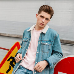 Wall Mural - Serious fashion pretty young man with hairstyle in fashionable blue casual jeans clothes near road metal signs outdoors. Modern good-looking guy in trendy jeans wear posing outdoors. Youth style.