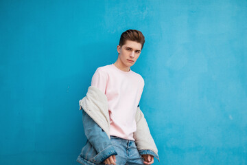 Wall Mural - Attractive young man with hairstyle in stylish pink sweatshirt in fashionable denim jacket posing near vintage colored wall in street. European handsome guy in jeans clothes is stand blue background.