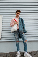 Wall Mural - Pretty fashionable young man in pink sweatshirt in denim jacket in ripped jeans with fabric bag posing near vintage building in city on summer day. Attractive guy in casual wear with shopper outdoors.