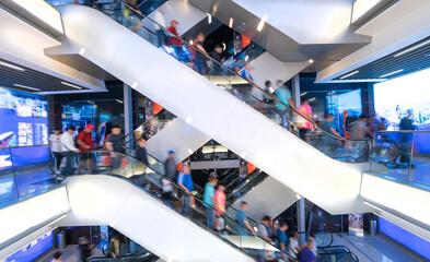 Customers clients moving on escalator staircases inside of giant modern shopping mall. Consumption concept.