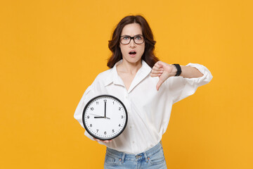 Wall Mural - Perplexed young brunette business woman in white shirt glasses isolated on yellow wall background studio. Achievement career wealth business concept. Mock up copy space. Hold clock showing thumb down.