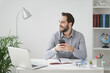 © ViDi Studio - Cheerful young business man in gray shirt sitting at desk work on laptop pc computer in light office on white wall background. Achievement business career concept. Using mobile phone, looking aside.