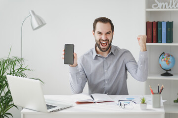 Wall Mural - Happy business man in gray shirt sit at desk work on laptop in light office on white wall background. Achievement business career concept. Hold mobile phone with blank screen doing winner gesture.