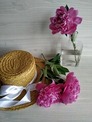 Two peonies lay on a wicker straw hat on a light wooden background. one peony is standing in a transparent vase. view from above