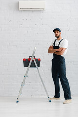 Wall Mural - Handsome workman in overalls looking at camera near toolbox on ladder and air conditioner on white wall