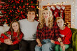 © Alina - Young happy family of four in Christmas clothes sitting and smiling in living room against Christmas lights.