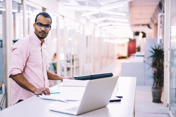 Portrait of handsome male engineer in trendy eyewear working in modern office on planning map of city infrastructure using official data from internet via laptop computer and fast 5G wireless