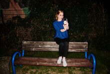 Portrait Of Young Woman Taking Selfie While Sitting On Bench In Park At Night