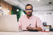 © BullRun - Portrait of confident and serious afro american businessman in spectacles looking at camera while writing some information in notepad during working process sitting in coworking space interior