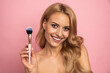 © Roman - Beauty portrait of a lovely young woman with long blonde hair standing isolated over pink background, holding makeup brush
