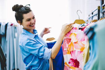 Happy female designer choosing dresses for own new collection standing in showroom with colorful clothing in hands.Fashion stylist working with production of trendy textile for development busines