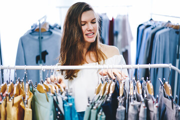 Wall Mural - Portrait of funny hipster girl enjoying shopping in mall choosing clothes on hanger during free time, beautiful woman working in trendy boutique selling fashionable productions of talented designers