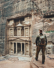 A Traveler In A Hat With A Backpack Looks At Al-Khazneh In Petra. Indiana Jones Near The Treasury In Petra. The Best View Of The Main Temple Of Al-Khazneh In Petra, Jordan.