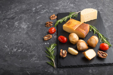 Smoked cheese and various types of cheese with rosemary and tomatoes on black slate board on a black concrete background. Side view, copy space