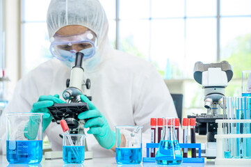 Lab equipment with blue liquid inside stand on the table with  male scientist wearing protection suit working with Microscope for research Coronavirus  vaccine at laboratory at the background.