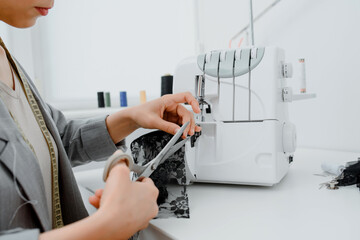 Wall Mural - Young woman tailor is cutting with scissors a piece of black material in her white mini workshop.