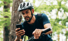 Professional Cyclist Resting In The Forest, Holding Mobile Phone, Using Online Application For Searching GPS Coordinates While Riding The Bike. Travel, Sport And Modern Technology Concept.