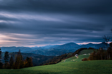 Idyllic green landscape in Slovenia. Hilltop covered with grass meadow and colorful forest trees. Dark clouds in the sky. Countryside dirt road with few houses. Amazing view of mountains in distance