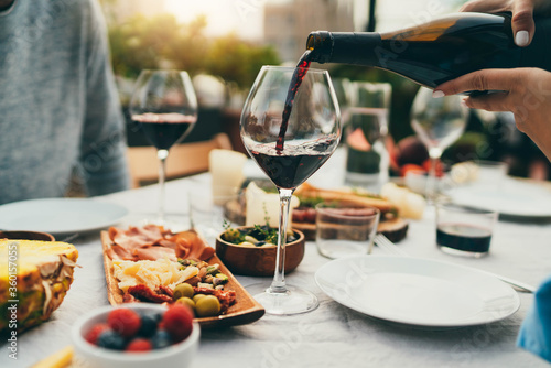 Outdoor terrace dinner with friends, woman pouring red wine in glass, Best friends drinking and eating delicious food on terrace, Young couple having romantic dinner at restaurant with healthy food