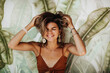 © Look! - Cheerful young woman with beautiful tan smiles widely and closes eyes. Portrait of emotional happy girl in brown top laughing on background of palm leafs