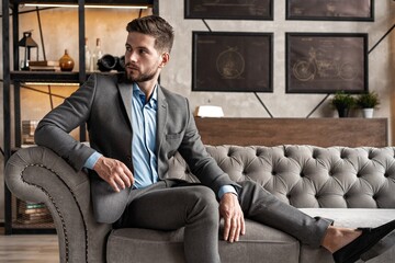 Wall Mural - Cool and handsome. Good looking young man in full suit sitting on the sofa while spending time indoors.