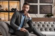 © opolja - Cool and handsome. Good looking young man in full suit sitting on the sofa while spending time indoors.