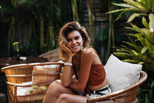 Cheerful Woman In Brown Top And Denim Shorts Smiles In Street Cafe. Tanned Girl In Summer Outfit Chilling Out And Sitting On Wooden Chair In Tropical Garden