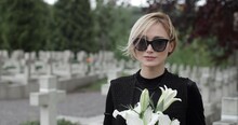 Close Up Of Woman In Dark Glasses Raising Head And Looking To Camera. Young Widow Holding White Lily Flower While Standing At Cemetery. Concept Of Memorial Day. Blurred Background