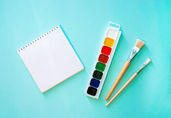 Watercolor paint with white, yellow, red, green, blue and black colors, two light beige brushes with white pile and wooden texture and white clear sheet of note book on a light blue background