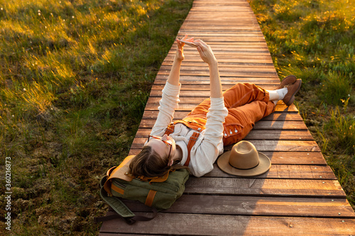 Fotomural Woman naturalist orange overalls makes selfie on the phone, resting lying on backpack on wooden path through peat bog swamp in wildlife national park at sunset