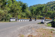 Bridge With Yellow Metal Railing Over River In Mountains On Bali Island. Decorative Stone Statues. People Ride Motorbikes. Passengers On The Means Of Transport. In The Background Tropical Vegetation