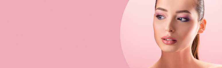 portrait of beautiful naked woman with shiny makeup looking through round paper hole isolated on pink, panoramic shot