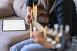 © Alvaro Sanchez/ADDICTIVE STOCK - Woman playing guitar sitting on her couch at home and learning with online lessons with a digital tablet with a digital tablet with a blank screen from above