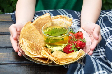 close-up of a man's hands holding a plate of pancakes with saucer of cream and strawberries.next to it is a transparent Cup of tea with blackcurrant and mint leaves on the background of wooden table