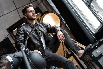 Wall Mural - Handsome young man in leather jacket looking away while sitting on the stool