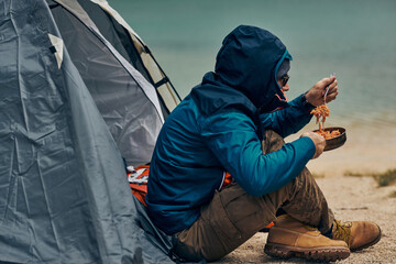 Man sitting on shore by the lake and eating his lunch on camping trip.