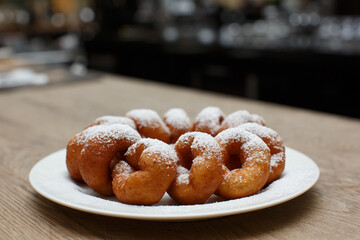 a pile of delicious sweet golden brown doughnuts (donuts) with white powdered icing sugar topping laying in a white plate on a wooden table background