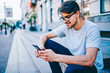 Cheerful male bogger reading funny comments from followers on website on smartphone with 4G internet.Positive hipster guy in eyewear reading notification with good new on telephone sitting in downtown